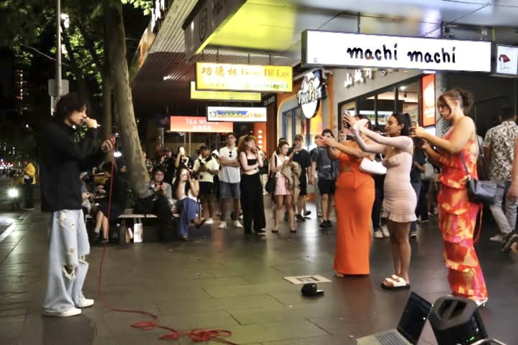 A wider Melbourne street scene with people watching KUMA perform outside the shops.