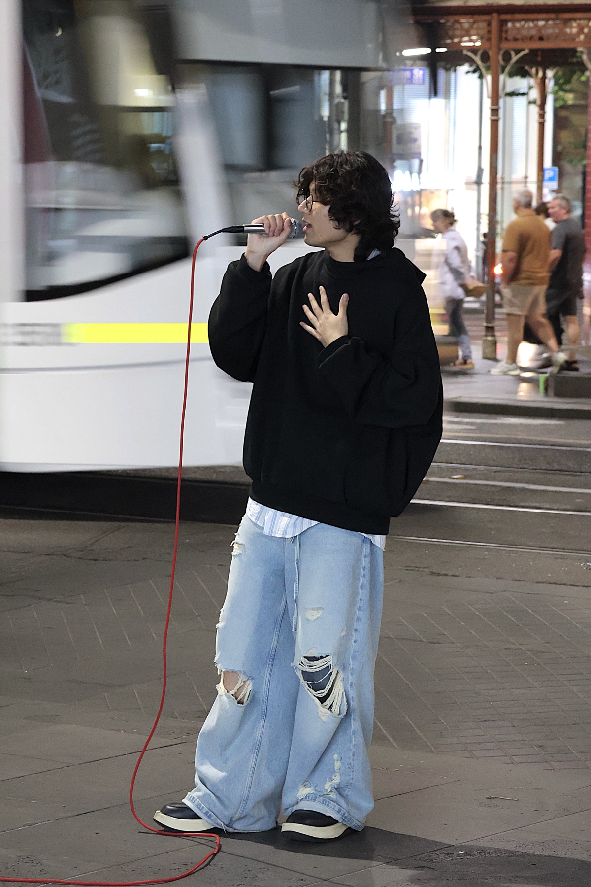 KUMA busking beside a passing Melbourne tram, wearing a black hoodie and light jeans.