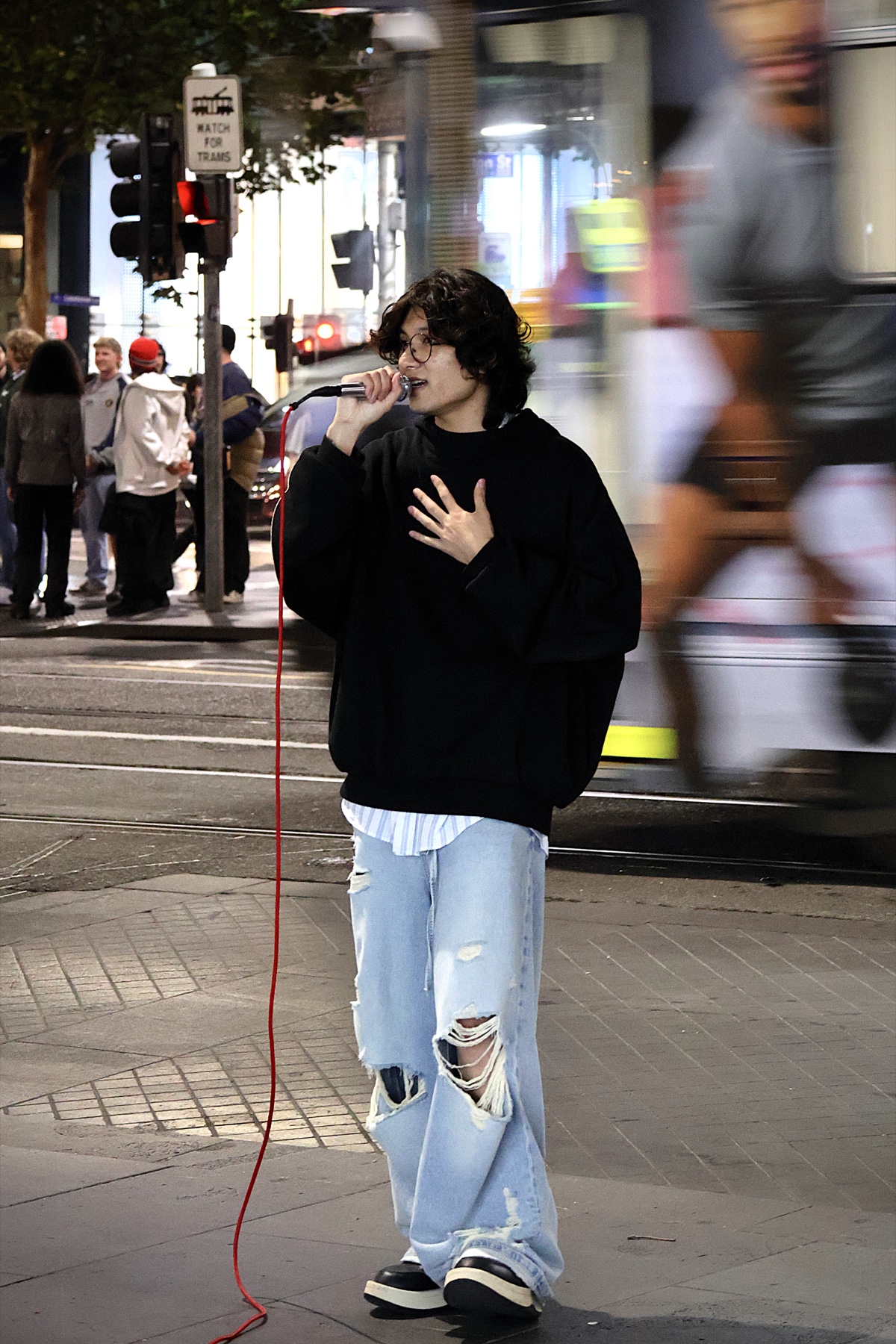 A closer portrait of KUMA singing on a busy Melbourne street as a tram passes behind him.