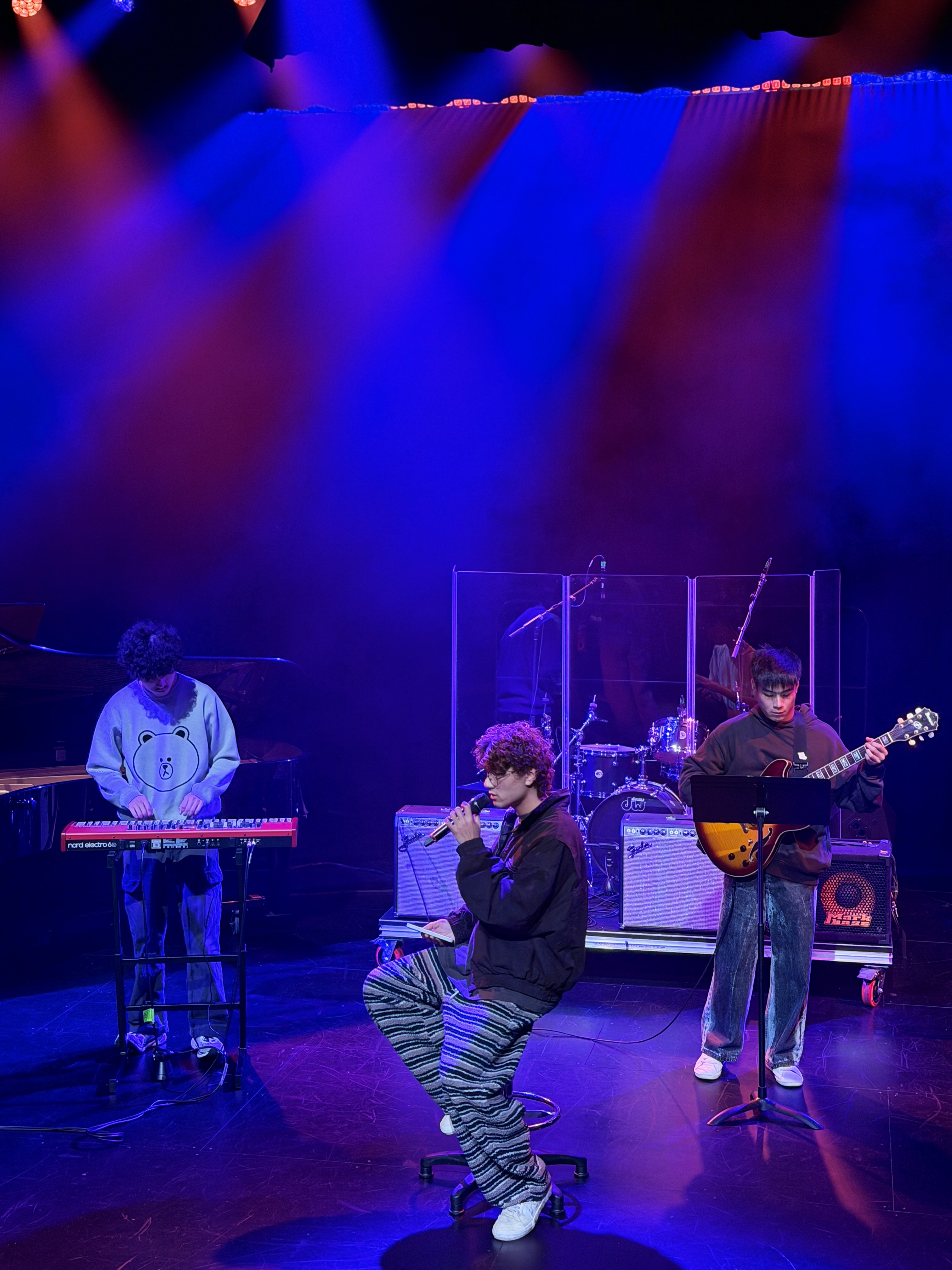KUMA singing with a keyboardist and guitarist under blue and red lights.