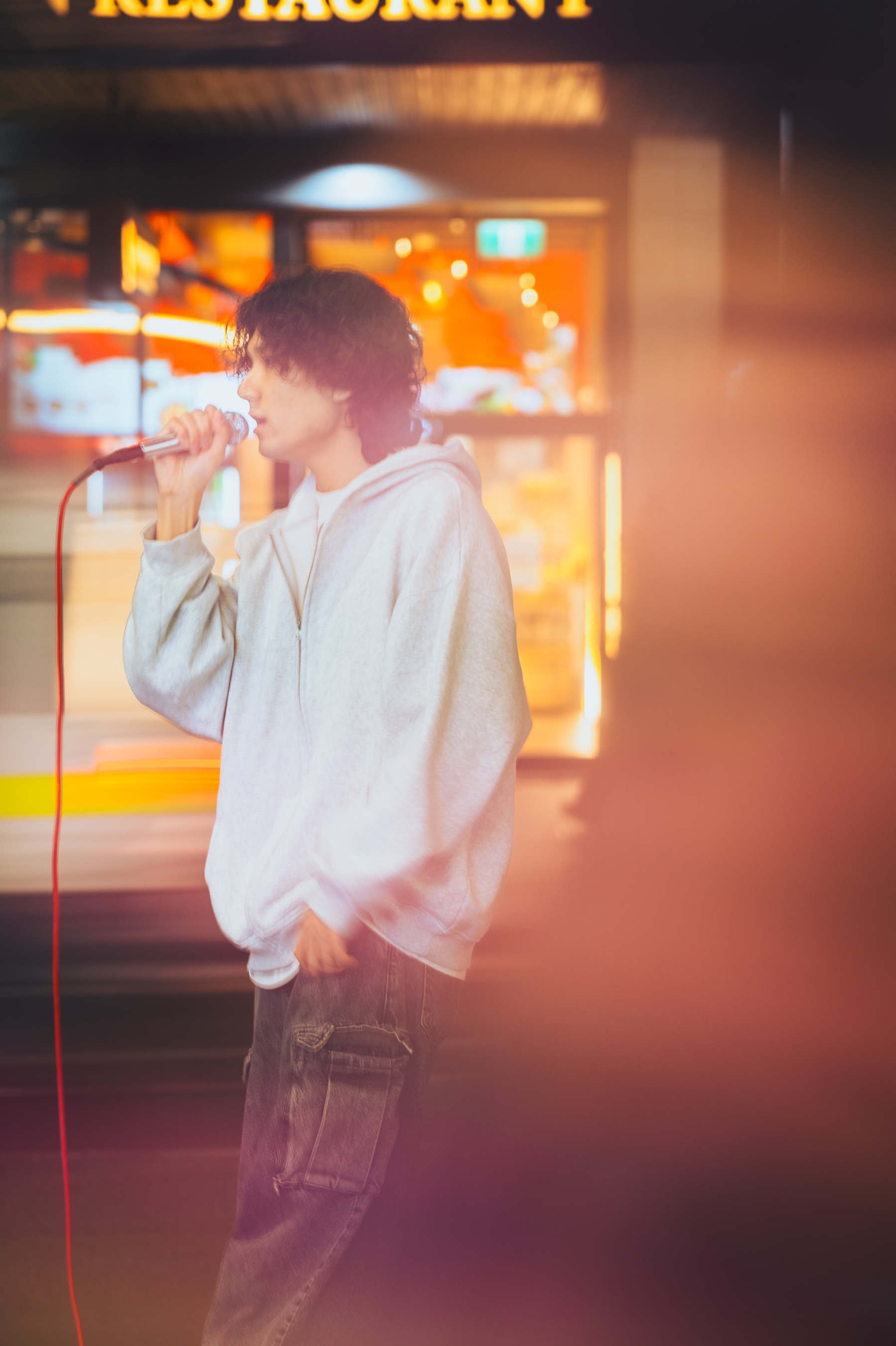 KUMA singing on a Melbourne street in a light hoodie, photographed in warm evening light.