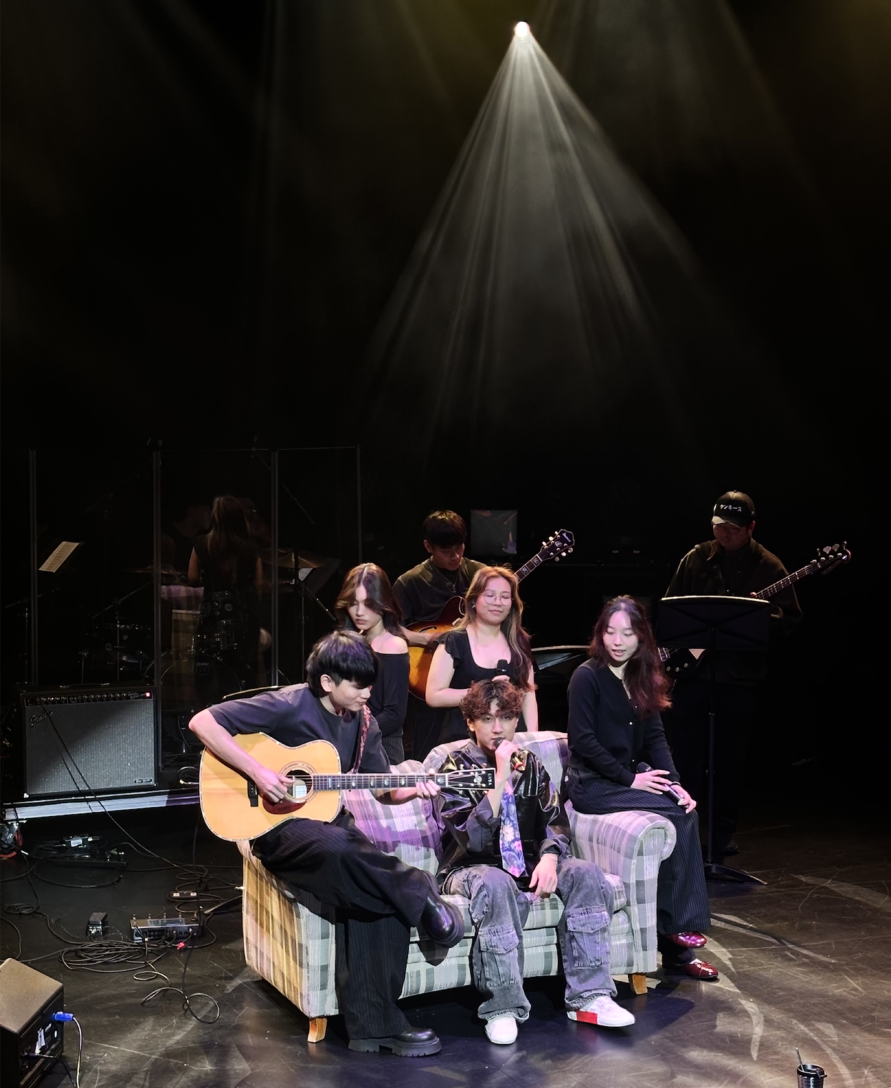 KUMA seated with an acoustic guitarist and fellow performers beneath a single spotlight.