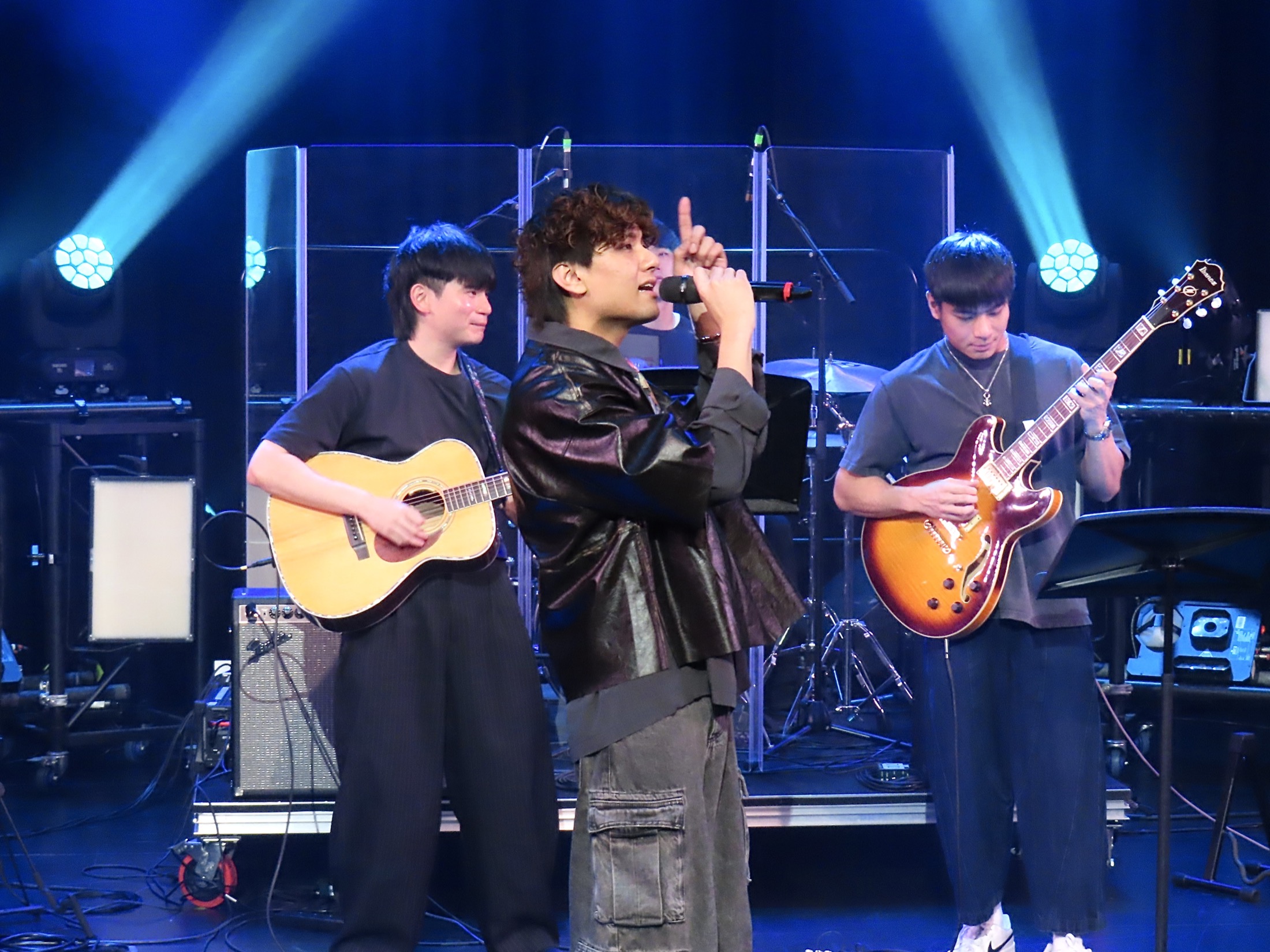 A wider alternate view of KUMA singing between two guitarists under blue stage lights.